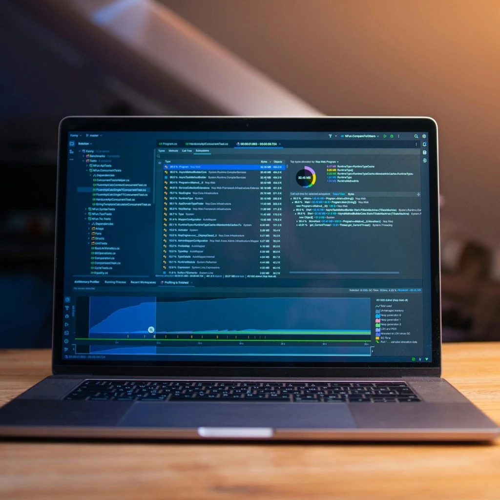 Laptop auf einem Holzschreibtisch zeigt eine professionelle Entwicklungsumgebung mit Code, Performance-Profiler und Live-Speicheranalyse im Dark-Mode — maßgeschneiderte Softwareentwicklung in Aktion.